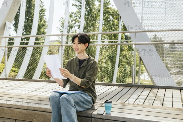 A young man study outside on wooden steps. The student smiling and reading notes. The atmosphere is casual, ideal for motivation and study