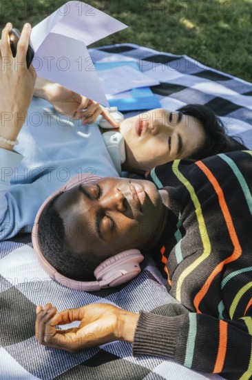 Friends relax outdoors on a sunny day, lying on a blanket. One listens to music with headphones, while the other reads under a clear sky, enjoying leisure time