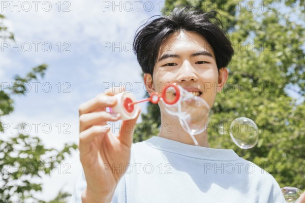 A smiling man enjoys blowing soap bubbles in a sunlit park. The clear sky and vibrant greenery create a cheerful and relaxing atmosphere, capturing a moment of lighthearted joy