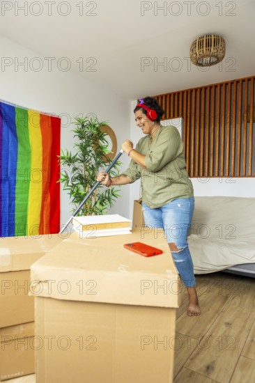 A trans woman smiling while organizing her new home. Surrounded by moving boxes and a pride flag, she listens to music with headphones in a welcoming environment