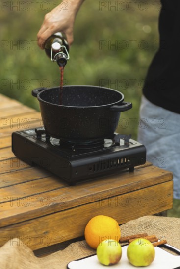 Cropped unrecognizable person pours red wine into a pot for making mulled wine outdoors, surrounded by fresh fruits and spices