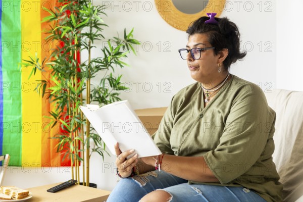 A trans woman enjoys a serene moment, sitting comfortably in her new home, reading a book. The background showcases a rainbow flag, symbolizing LGBTQIA+ pride and identity
