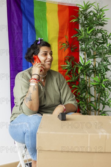 Trans woman sitting by moving boxes, talking on phone, smiling. Vibrant rainbow pride flag in background, embracing identity and new beginnings at home