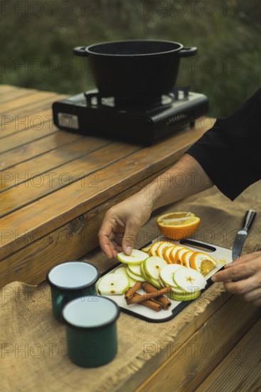 Cropped unrecognizable person preparing ingredients for mulled wine outdoors, arranging sliced apples and oranges on a wooden table