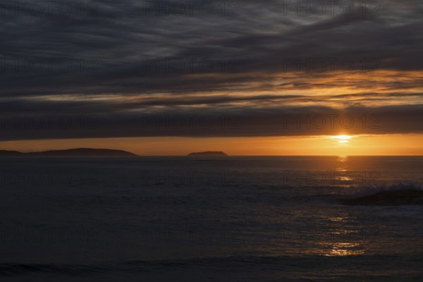 A serene sunset over the ocean, with the sun casting radiant orange and yellow hues across the sky. Silhouetted islands in the distance add to the tranquil beauty, with calm waters reflecting the vibrant colors amidst the dark, cloudy sky