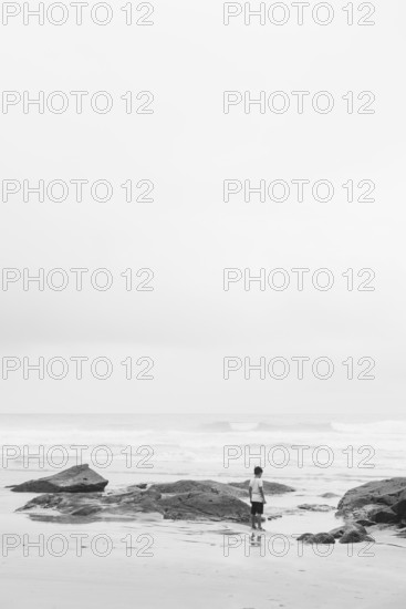 A lone individual stands on a beach surrounded by rocks, gazing at the ocean. The black and white photograph captures a sense of solitude, tranquility, and reflection in an expansive, minimalist setting