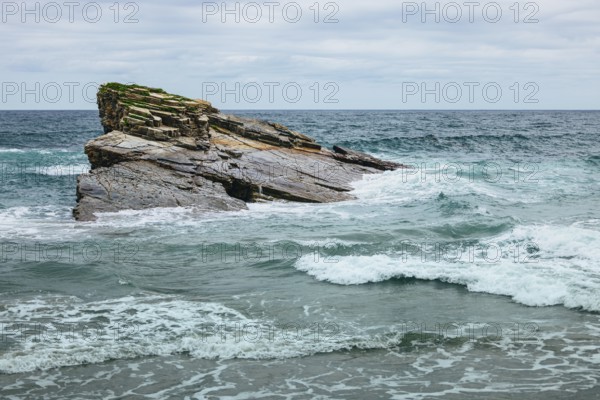 A scenic view of a rocky cliff surrounded by ocean waves crashing under a cloudy sky. The intricate rock formations and the dynamic movement of the waves create a captivating and serene coastal landscape