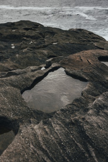 A serene coastal scene features rugged rock formations with shallow pools of water collected within the crevices. The ocean waves create a tranquil, yet dynamic backdrop, highlighting the natural beauty of the shoreline