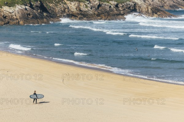 A surfer carrying a surfboard walks alone on a sandy beach toward the ocean, with waves crashing against rocky cliffs in the background. The beach is deserted, creating a sense of peaceful solitude and connection with nature