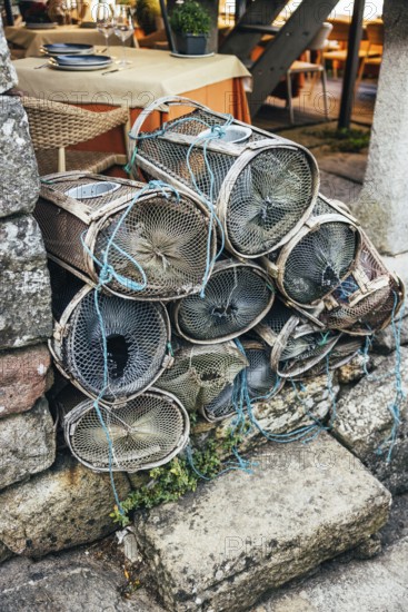 A stack of fishing traps rests against a rough stone wall beside an outdoor cafe table. The scene captures the rustic charm of coastal life with wicker chairs and a table set for dining nearby