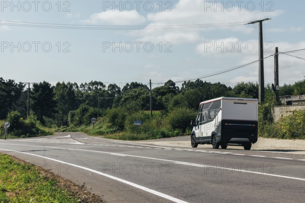 A motorhome drives on a serene rural road surrounded by lush greenery and power lines under a clear sky. The landscape offers a peaceful and inviting atmosphere for a road trip or outdoor adventure