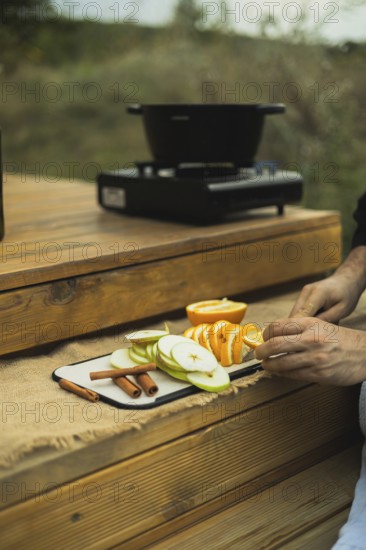 Cropped, unrecognizable person slices fresh oranges and apples, arranging them with cinnamon sticks outdoors, prepping for homemade mulled wine
