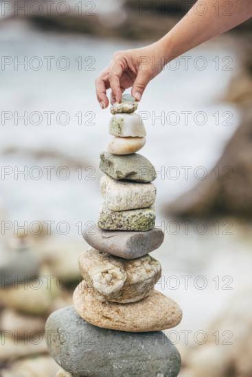 A hand carefully places a small stone atop a balanced stack of larger rocks by the seaside. The focus is on the stones and the hand, with the sea softly blurred in the background, symbolizing balance and mindfulness