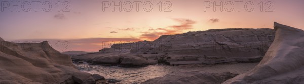Sweeping panoramic view of Sarakiniko beach on Milos Island, Greece, under a vibrant sunset sky. The unique white volcanic rock formations gracefully surround a tranquil sea