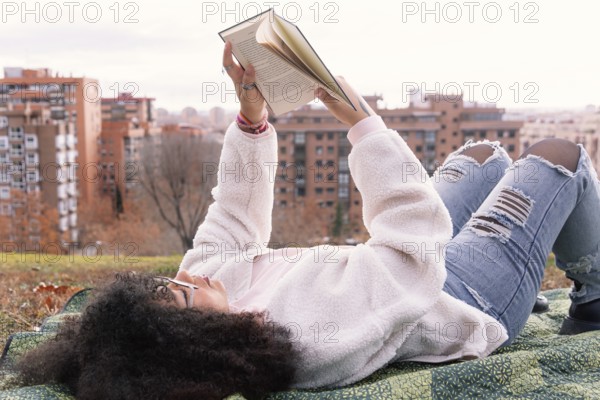 A woman lies on a blanket in a park, reading a book. They wear casual clothes, including jeans and a fluffy jacket. In the background, city buildings and autumn trees are visible