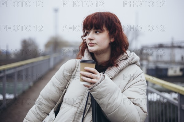 A young woman stands on a pedestrian bridge, wearing a warm jacket and holding a cup of coffee in a day of travel by train