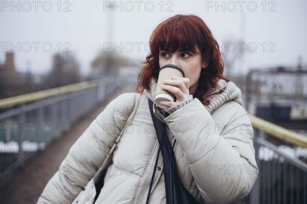A young woman in a warm winter coat drinks coffee from a disposable cup in a day of travel by train, standing on a pathway with a blurred background