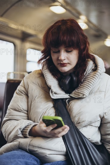 A young woman in a cozy coat, with headphones around her neck, focuses on her smartphone while traveling on a train, showcasing a moment of digital connectivity in transit