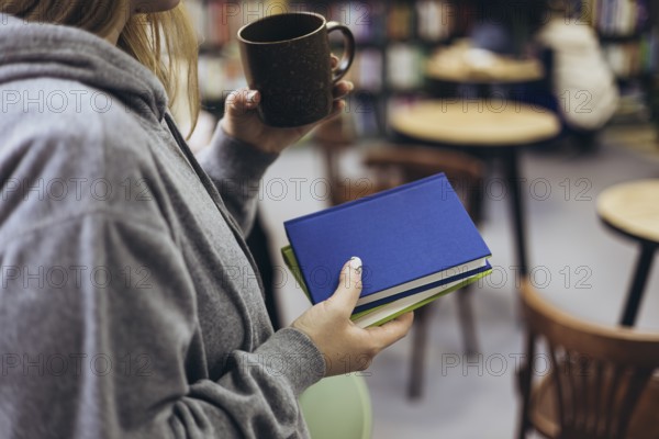 A woman in a gray hoodie holds books and a cup of coffee, enjoying a relaxed moment in a cozy bookstore. Shelves and chairs add to the inviting atmosphere