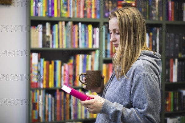 A woman in a gray hoodie enjoys a quiet moment in a bookstore. They hold a pink book and a brown mug, surrounded by colorful books on shelves
