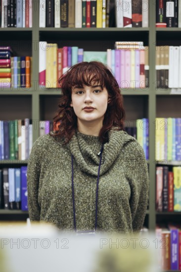 A young female academic pauses thoughtfully in a library, surrounded by colorful bookshelves filled with numerous books, showcasing a contemplative expression