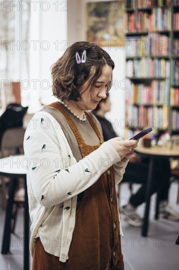 A young woman with a casual style checks her phone in a cozy bookstore. Surrounded by vibrant bookshelves, she seems focused and relaxed, enjoying her leisure time
