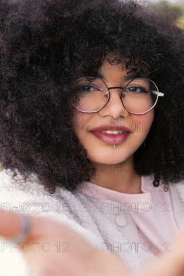 A young latin woman with voluminous curly hair, wearing round glasses and a cozy sweater, smiles warmly. Her facial piercings add a modern edge to her casual style and friendly expression