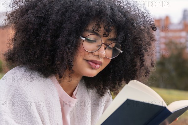 A young latin woman with curly hair and glasses reads a book outdoors, enjoying a peaceful and relaxing environment. She is dressed casually and appears content and focused on her reading