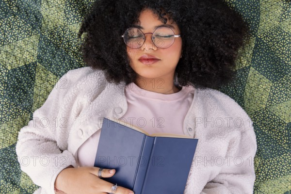 A latin woman with curly hair and glasses peacefully rests on a patterned blanket, holding a book. She appears relaxed and content, enjoying a quiet moment