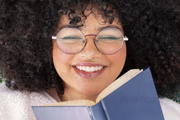 A joyful latin woman with curly hair and large round glasses, holding a book with delight. Her bright smile and casual attire enhance the positive, relaxed vibe