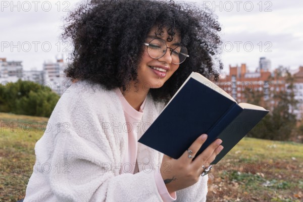 A latin woman with curly hair and glasses enjoys reading a book in an outdoor park setting. She is sitting on the grass, looking relaxed and content, with a cityscape in the background