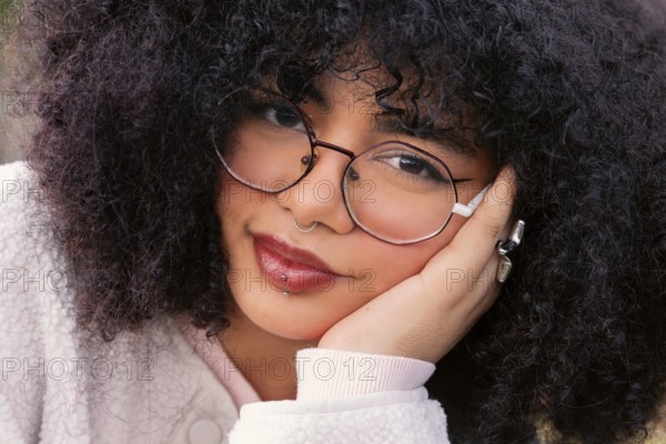 A young woman with curly hair wearing glasses and piercings is captured up-close. Her thoughtful expression reflects confidence and a modern sense of style