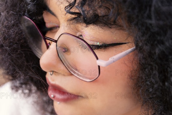 A detailed close-up shot of a latin woman wearing glasses. The focus highlights their piercings, makeup, and curly hair, capturing a unique style and expression in soft, natural light