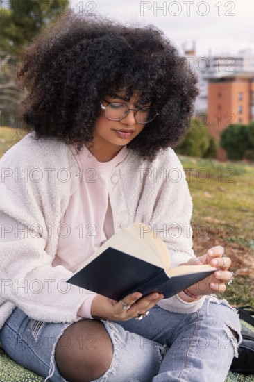 A latin woman with curly hair and glasses enjoys reading a book while seated on grass in a peaceful park. She is wearing a cozy sweater and ripped jeans, highlighting casual comfort
