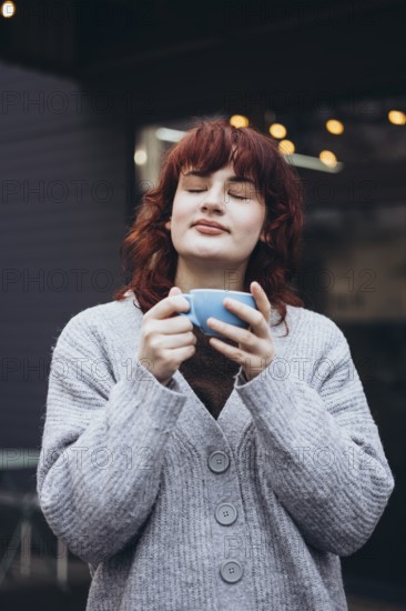 A young woman with closed eyes savors a moment with a cup of coffee. She wears a cozy gray sweater, standing outdoors on cafe terrace, creating a serene and content atmosphere