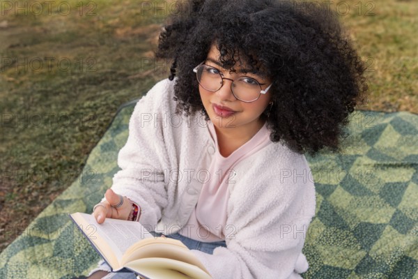 A young latin woman with curly hair and glasses sits on a picnic blanket outdoors, absorbed in reading a book. The serene setting suggests relaxation and leisure under a warm sun