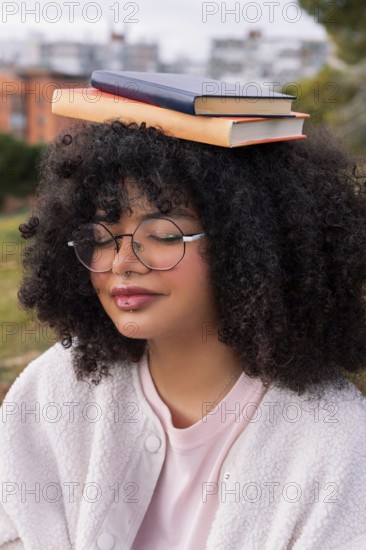 A young latin woman with curly hair and glasses balances two books on her head while outdoors in a park. She has a serene expression, embodying calmness and focus