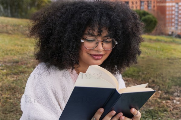 A young latin woman with curly hair and glasses reads a book while sitting on grass in a park. Her relaxed expression reflects her enjoyment of literature and nature
