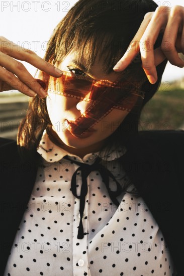 Woman in polka dot blouse, holding film strip against sunlight, creating an artistic shadow over her face. The sun enhances the warm tones and ethereal feel