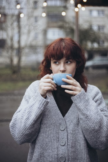 A young woman enjoys a coffee from a blue cup while standing outdoors on cafe terrace. She is wearing a cozy gray cardigan, creating a serene atmosphere under soft lights