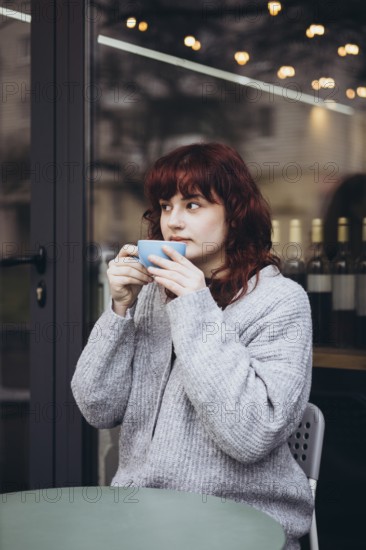 A young woman in a cozy sweater enjoys her coffee on a cafe terrace. The ambiance is warm, with a calm, contemplative mood, enhanced by soft lighting and reflections