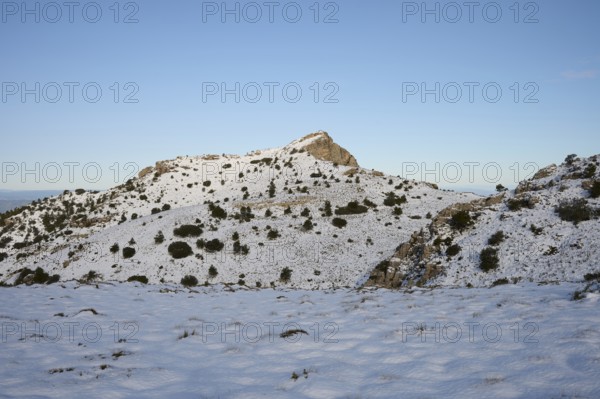 A serene winter scene at Montcabrer in Spain, showcasing its snow-capped peak under a clear blue sky. Sparse vegetation dots the landscape, adding to the picturesque view