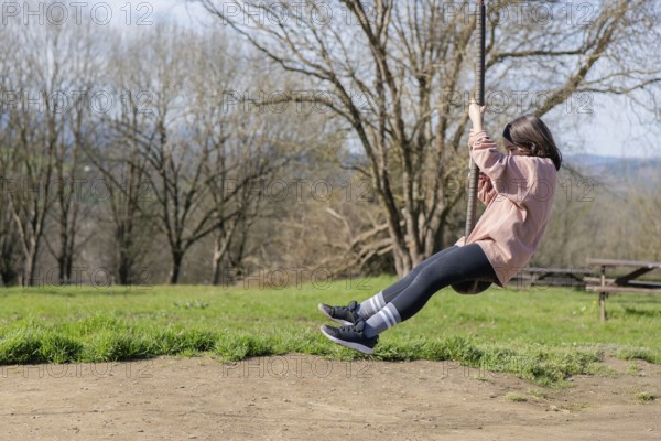 A young girl enjoys a whimsical ride on a zip line in a sunny park. Her casual attire and the vibrant green landscape create a sense of joy and freedom