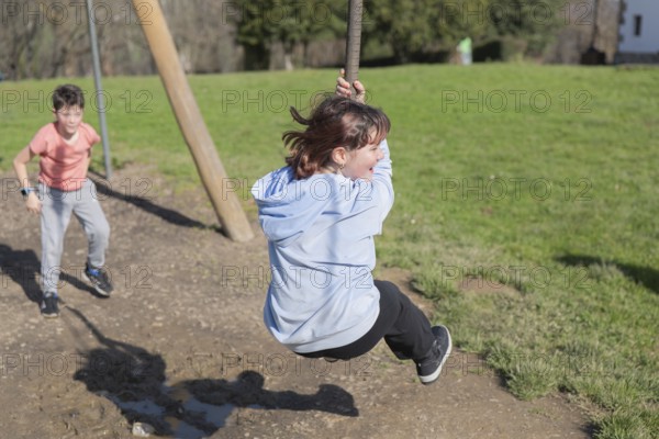 Two friends enjoy playing on a zip line in a sunny park. One child swings while the other waits in anticipation. The scene captures excitement and outdoor fun