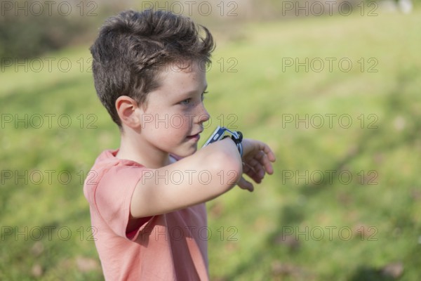 A young boy in a coral shirt examines his smartwatch in a sunny park. The background features blurred greenery, capturing a sense of exploration and curiosity