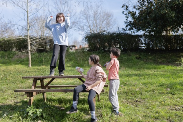 Three friends playfully interact on a wooden picnic table in a sunny park. One child stands energetically on the table, while the others watch and enjoy their time