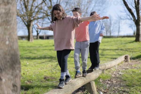 Three friends are balancing on a wooden beam in a park. They are enjoying the sunny day, surrounded by green grass and trees, showcasing friendship and playfulness