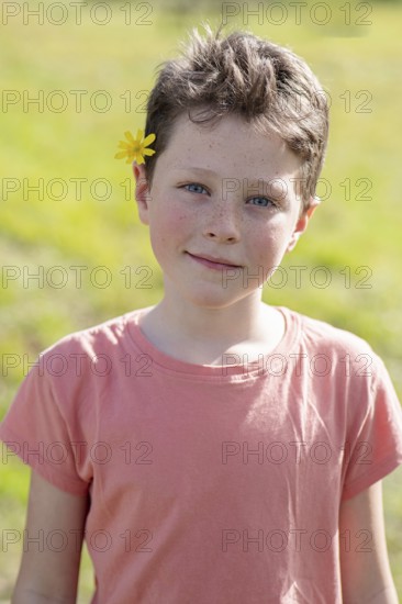 A young child with short hair and a flower tucked behind the ear enjoys a sunny day outdoors. The child is wearing a casual pink shirt, smiling subtly, and surrounded by greenery