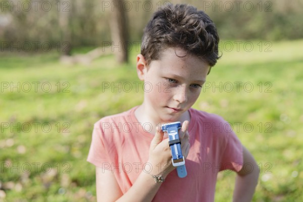 Young boy in pink shirt holding a blue smartwatch in a green park. Sunlit scene with blurred background, conveying curiosity and playfulness