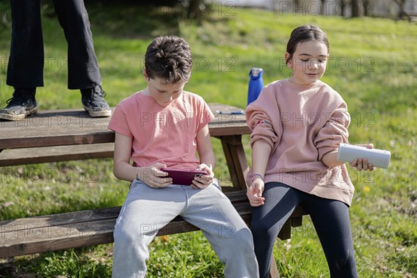 Two friends sit on a wooden bench outdoors, immersed in technology. One uses a tablet, while the other holds a portable speaker, enjoying a warm day in nature